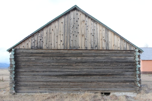 A rustic wooden barn with a mix of horizontal logs and vertical planks dominates the scene. Weathered brown logs form the lower section, while the upper section consists of lighter, vertical wooden planks arranged in a triangular shape, suggesting a gable roof. The barn is set in a field with dry grass, and a partially visible structure with a reddish wall is seen to the side.