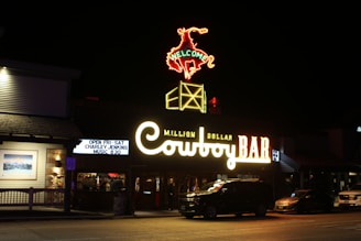A night scene featuring a brightly illuminated sign for the 'Million Dollar Cowboy Bar'. The neon sign includes a cowboy riding a bucking bronco with 'Welcome' written above. Below, the bar's name is prominently displayed in bright yellow and orange lights. A few cars are parked in front, and there's a sidewalk and storefront visible as well.