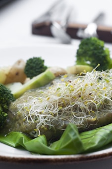 A plate featuring a dish garnished with alfalfa sprouts, accompanied by vegetables including broccoli, zucchini slices, and baby potatoes. The vegetables are arranged around a sauce-covered main component. The background includes blurred cutlery.
