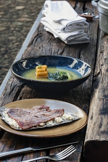 A rustic wooden table is set with two dishes. The first dish contains a blue bowl filled with soup, featuring corn and greens. The second dish has a plate with a piece of grilled meat served on a flatbread with crumbled cheese. White napkins are neatly stacked beside these dishes.