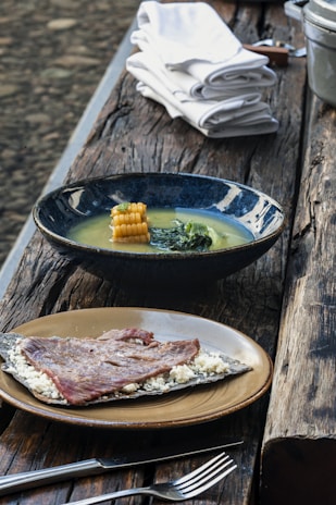 A rustic wooden table is set with two dishes. The first dish contains a blue bowl filled with soup, featuring corn and greens. The second dish has a plate with a piece of grilled meat served on a flatbread with crumbled cheese. White napkins are neatly stacked beside these dishes.