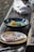 A rustic wooden table is set with two dishes. The first dish contains a blue bowl filled with soup, featuring corn and greens. The second dish has a plate with a piece of grilled meat served on a flatbread with crumbled cheese. White napkins are neatly stacked beside these dishes.