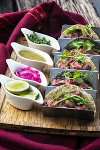 A wooden board features a set of three tacos filled with cooked meat and topped with fresh greens. Accompanying the tacos are small ceramic dishes containing various condiments, such as chopped herbs, pickled onions, lime wedges, and a green sauce. A vibrant red fabric is draped around the setup, adding a pop of color.