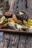 A wooden board is laid with a variety of traditional Mexican foods, including corn tortillas topped with refried beans and cheese, fried empanadas, avocado slices, lime wedges, and small bowls of salsa and bean paste. The arrangement includes roasted vegetables and garnishes of green herbs. The board sits on a rustic wooden surface, suggesting an outdoor setting.
