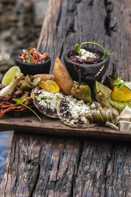 A wooden board is laid with a variety of traditional Mexican foods, including corn tortillas topped with refried beans and cheese, fried empanadas, avocado slices, lime wedges, and small bowls of salsa and bean paste. The arrangement includes roasted vegetables and garnishes of green herbs. The board sits on a rustic wooden surface, suggesting an outdoor setting.