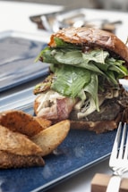A gourmet burger is presented on a blue plate. The burger is stacked with fresh greens, bacon, and a juicy patty, all held together with a glossy brioche bun. Next to the burger, there are seasoned potato wedges. A fork and knife are partially visible on the side.