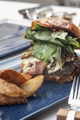 A gourmet burger is presented on a blue plate. The burger is stacked with fresh greens, bacon, and a juicy patty, all held together with a glossy brioche bun. Next to the burger, there are seasoned potato wedges. A fork and knife are partially visible on the side.
