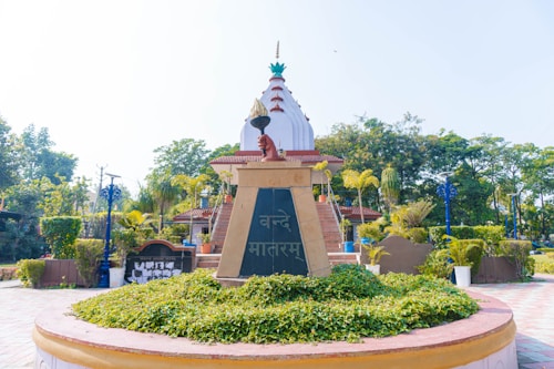 A monument featuring a decorative dome-like structure with intricate designs is surrounded by lush green plants and trees. In front of the monument, there is a pedestal with a Hindi inscription and a sculpture or emblem on top. The area is well-maintained with potted plants and neatly arranged bushes. The clear blue sky enhances the overall serene and peaceful atmosphere.