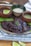 A dish featuring succulent meat covered in a rich, dark sauce garnished with fresh herbs. It is presented on a plate that rests on a textured cloth, with slices of lime nearby. The scene includes small bowls containing sauce and rice, suggesting an appetizing, well-rounded meal.