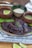 A dish featuring succulent meat covered in a rich, dark sauce garnished with fresh herbs. It is presented on a plate that rests on a textured cloth, with slices of lime nearby. The scene includes small bowls containing sauce and rice, suggesting an appetizing, well-rounded meal.