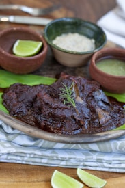 A dish featuring succulent meat covered in a rich, dark sauce garnished with fresh herbs. It is presented on a plate that rests on a textured cloth, with slices of lime nearby. The scene includes small bowls containing sauce and rice, suggesting an appetizing, well-rounded meal.