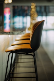 A row of four modern bar stools with orange seats and black backs, aligned in front of a counter with a blurred background featuring colorful lights and bokeh effects, likely in an upscale or stylish bar, restaurant, or cafe.