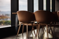 A set of modern wooden chairs with curved backs arranged in a row near large windows, allowing natural light to illuminate the space. The chairs and the floor have a warm wooden finish, and the windows offer a blurred view of an urban landscape outside.