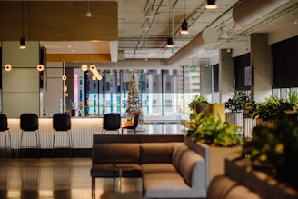 A modern, stylish lounge area featuring a bar with high chairs, decorative hanging lights, and indoor plants. There's a cozy seating arrangement with sofas and a small, decorated Christmas tree in the background. The space is well lit, with large windows allowing natural light to enter.