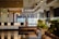 A modern, stylish lounge area featuring a bar with high chairs, decorative hanging lights, and indoor plants. There's a cozy seating arrangement with sofas and a small, decorated Christmas tree in the background. The space is well lit, with large windows allowing natural light to enter.