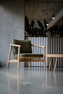 A minimalist and modern chair with wooden legs and a green cushion is placed on a sleek, polished concrete floor. In the background, there is a large, metallic plant container with lush green foliage, under artificial ceiling lighting.