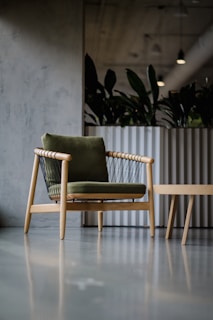 A minimalist and modern chair with wooden legs and a green cushion is placed on a sleek, polished concrete floor. In the background, there is a large, metallic plant container with lush green foliage, under artificial ceiling lighting.