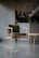 A minimalist and modern chair with wooden legs and a green cushion is placed on a sleek, polished concrete floor. In the background, there is a large, metallic plant container with lush green foliage, under artificial ceiling lighting.
