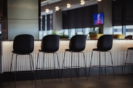 A modern bar setting with high black chairs positioned in front of a counter. The backdrop includes soft lighting and contemporary design elements. In the background, a television screen is mounted on the wall, slightly blurred.