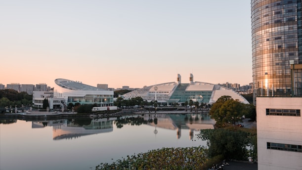 A modern landscape featuring futuristic architecture with sleek curves and reflective glass surfaces. The buildings are situated beside a tranquil body of water, with an urban skyline visible in the background. The sunset casts a warm glow, reflecting on the glass and water, creating a picturesque and serene scene.