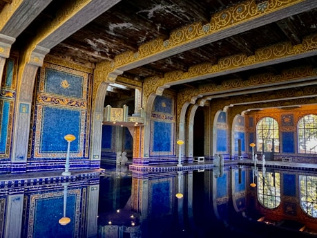 An opulent indoor pool with intricate blue and gold mosaic tiles covers the walls and ceiling, reflecting beautifully in the still water below. The architecture is ornate, featuring arches and columns. Soft lighting enhances the luxurious and serene atmosphere.