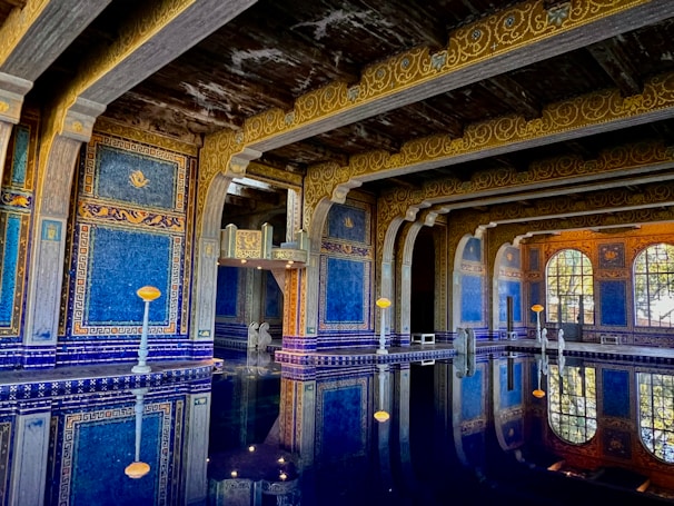 An opulent indoor pool with intricate blue and gold mosaic tiles covers the walls and ceiling, reflecting beautifully in the still water below. The architecture is ornate, featuring arches and columns. Soft lighting enhances the luxurious and serene atmosphere.