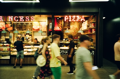 A bustling food stall with pizzas on display, where several people are walking by. The interior has a warm ambiance with glowing signage and decorative elements. The counter area is filled with various products and ingredients, adding to the busy atmosphere.