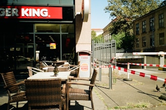A street view of a Burger King restaurant with outdoor seating featuring woven chairs and tables. A bird is perched on one of the tables. The surroundings include some barricades with red and white tape and a building entrance with trees in the background.