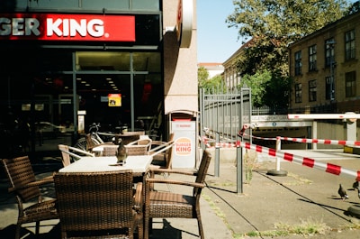 A street view of a Burger King restaurant with outdoor seating featuring woven chairs and tables. A bird is perched on one of the tables. The surroundings include some barricades with red and white tape and a building entrance with trees in the background.