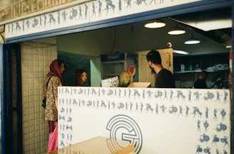 A small shop or cafe with a counter designed with blue silhouettes of figures in various poses against a white background. Three people are inside the establishment, engaged in conversation. One wears a headscarf, another has long dark hair, and the third is looking down at a counter or surface. The interior features subdued lighting with hanging lamps. A Pepsi fridge is visible in the background.