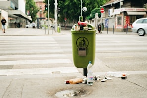 A green trash can overflowing with various types of litter, including bottles and cans, is situated on a city sidewalk. The surrounding area shows a crosswalk with some pedestrians in the distance. Nearby shops and parked cars create an urban environment, with visible greenery in the background.