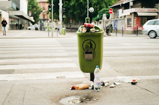A green trash can overflowing with various types of litter, including bottles and cans, is situated on a city sidewalk. The surrounding area shows a crosswalk with some pedestrians in the distance. Nearby shops and parked cars create an urban environment, with visible greenery in the background.