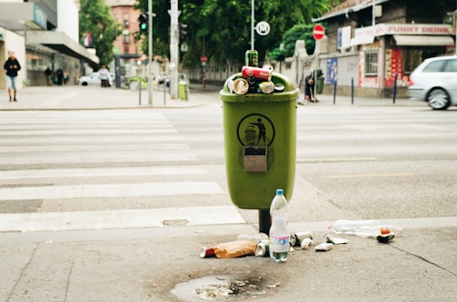 A green trash can overflowing with various types of litter, including bottles and cans, is situated on a city sidewalk. The surrounding area shows a crosswalk with some pedestrians in the distance. Nearby shops and parked cars create an urban environment, with visible greenery in the background.