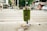 A green trash can overflowing with various types of litter, including bottles and cans, is situated on a city sidewalk. The surrounding area shows a crosswalk with some pedestrians in the distance. Nearby shops and parked cars create an urban environment, with visible greenery in the background.
