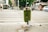 A green trash can overflowing with various types of litter, including bottles and cans, is situated on a city sidewalk. The surrounding area shows a crosswalk with some pedestrians in the distance. Nearby shops and parked cars create an urban environment, with visible greenery in the background.