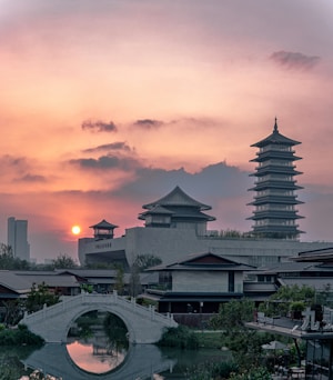 A serene landscape featuring traditional East Asian architecture, including a prominent multi-tiered pagoda and curved-roof buildings. A stone arch bridge crosses a calm river, and the setting sun casts a warm glow across the sky with scattered clouds.