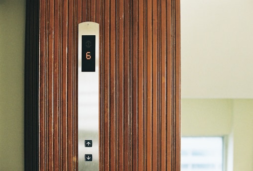A wooden elevator panel with vertical grooves features a modern display indicating the number 6 in illuminated digits. Below the display, there are buttons with arrows pointing up and down, set against a sleek metallic strip.