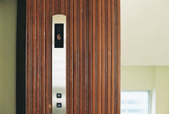 A wooden elevator panel with vertical grooves features a modern display indicating the number 6 in illuminated digits. Below the display, there are buttons with arrows pointing up and down, set against a sleek metallic strip.