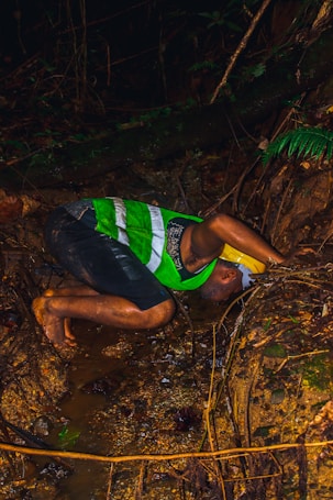 A person is crouched down, drinking water from a small stream in a forest environment. The individual is wearing a green and white striped shirt with dark shorts. The surroundings are dense with rich green foliage, rocks, and exposed roots.