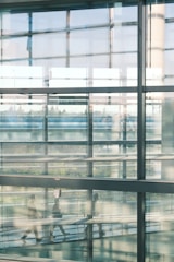 A modern architectural scene with large glass panels reflecting blurred silhouettes of people walking inside a building. The interior has an industrial feel with metal beams and a view of greenery visible through the windows.