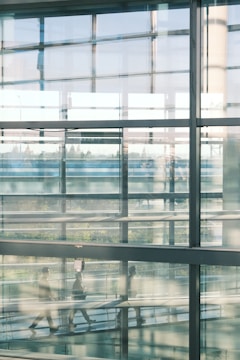 A modern architectural scene with large glass panels reflecting blurred silhouettes of people walking inside a building. The interior has an industrial feel with metal beams and a view of greenery visible through the windows.