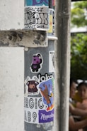 A metal pole covered with a variety of colorful stickers. These include cartoon characters, graffiti tags, and text with bold designs. The stickers appear to have been collected over time, showcasing playful and artistic themes. In the background, two people are vaguely visible, adding a touch of urban life to the scene.