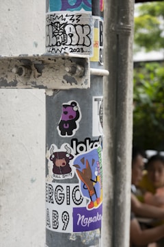 A metal pole covered with a variety of colorful stickers. These include cartoon characters, graffiti tags, and text with bold designs. The stickers appear to have been collected over time, showcasing playful and artistic themes. In the background, two people are vaguely visible, adding a touch of urban life to the scene.