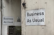 Two white signs with black text that read 'Business As Usual' are mounted on the exterior walls of a building. The walls appear weathered and slightly stained. One sign is on a larger wall, while the other is on a smaller, adjacent wall. There is also a small, triangular warning sign nearby and a metal utility box with an address written on it.