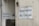Two white signs with black text that read 'Business As Usual' are mounted on the exterior walls of a building. The walls appear weathered and slightly stained. One sign is on a larger wall, while the other is on a smaller, adjacent wall. There is also a small, triangular warning sign nearby and a metal utility box with an address written on it.