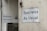 Two white signs with black text that read 'Business As Usual' are mounted on the exterior walls of a building. The walls appear weathered and slightly stained. One sign is on a larger wall, while the other is on a smaller, adjacent wall. There is also a small, triangular warning sign nearby and a metal utility box with an address written on it.