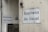 Two white signs with black text that read 'Business As Usual' are mounted on the exterior walls of a building. The walls appear weathered and slightly stained. One sign is on a larger wall, while the other is on a smaller, adjacent wall. There is also a small, triangular warning sign nearby and a metal utility box with an address written on it.