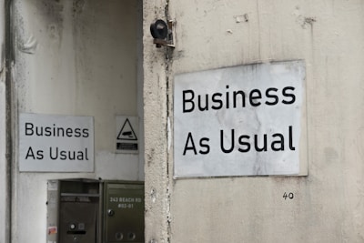 Two white signs with black text that read 'Business As Usual' are mounted on the exterior walls of a building. The walls appear weathered and slightly stained. One sign is on a larger wall, while the other is on a smaller, adjacent wall. There is also a small, triangular warning sign nearby and a metal utility box with an address written on it.