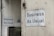 Two white signs with black text that read 'Business As Usual' are mounted on the exterior walls of a building. The walls appear weathered and slightly stained. One sign is on a larger wall, while the other is on a smaller, adjacent wall. There is also a small, triangular warning sign nearby and a metal utility box with an address written on it.