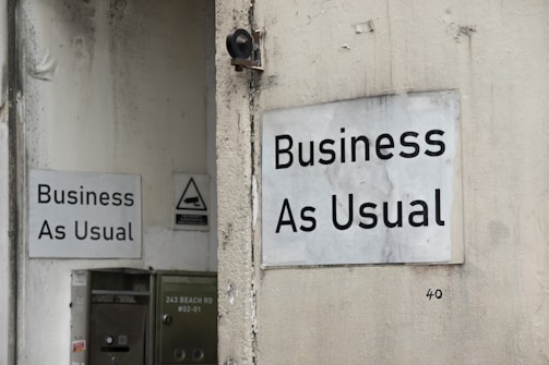 Two white signs with black text that read 'Business As Usual' are mounted on the exterior walls of a building. The walls appear weathered and slightly stained. One sign is on a larger wall, while the other is on a smaller, adjacent wall. There is also a small, triangular warning sign nearby and a metal utility box with an address written on it.
